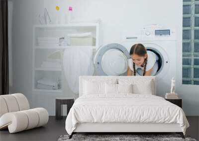 Pretty little girl householder poses inside of washing machine, takes checkered shirt from basin, engaged in laundry, has glad expression, two combed plaits. Childhood and washing day concept Wall mural