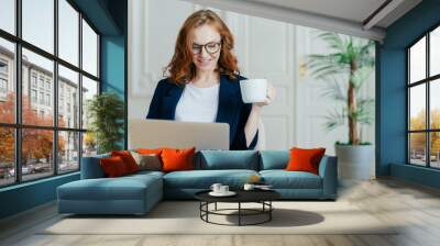 Satisfied European female with ginger hair, types information on laptop computer, has glad facial expression, drinks coffee, wears spectacles, develops new project, poses in cosy room alone. Wall mural