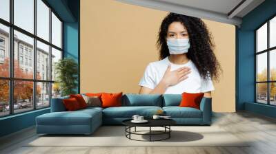 Serious Afro American woman wears medical face mask, has problems with breathing, presses hand to chest, got infected with coronavirus, isolated on beige ackground. Covid 19, health care concept Wall mural