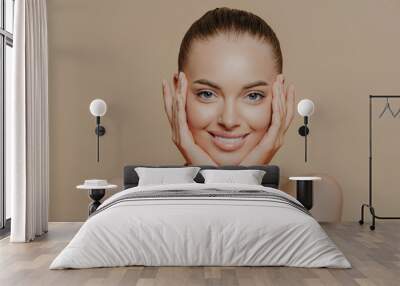 Skin care and beauty concept. Headshot of beautiful cheerful woman touches face gently, has perfect smile, healthy skin after cleaning or applying facial mask, isolated over beige background Wall mural