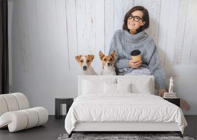 Smiling cheerful young female wears warm woolen sweater, square eyewear, drinks hot beverage, relaxes on floor and her two favourite pets, look directly into camera. People and animals conecpt Wall mural