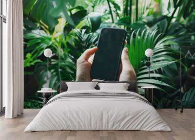 Someone holding a smartphone amidst lush green tropical plants, emphasizing the blend of technology and nature. Wall mural