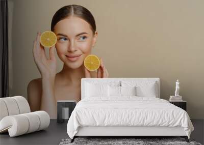 Thoughtful young European woman uses homemade fruit for facial mask holds lemon slices wrapped in white soft bath towel Wall mural