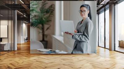 Woman administrative stands near desktop, holds modern laptop computer, uses wireless connection to internet, dressed in formal grey outfit, searches information on web page, works distantly Wall mural