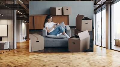 Woman in armchair clicks phone among boxes. European girl having rest in apartment after relocation. Wall mural