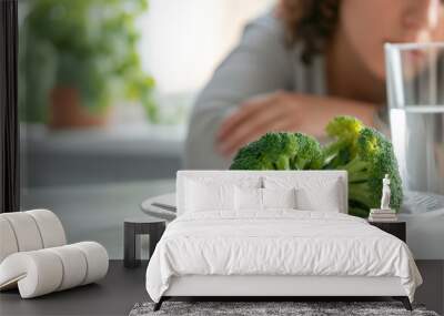 close-up of a single broccoli floret on a white plate, with a fork beside it, a glass of water in the background, and a person's hands folded in the background Wall mural