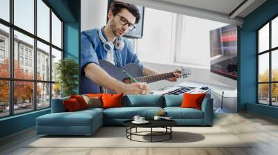 Male music arranger composing song on midi piano and audio equipment in digital recording studio. Man plays guitar and produce electronic soundtrack or track in project at home. Wall mural