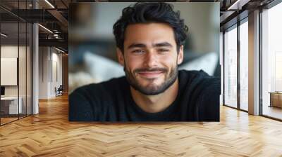 Smiling man with dark wavy hair wearing a black sweater, sitting in a cozy indoor space, relaxed and casual, warm atmosphere, confident expression, bright smile, candid portrait. Wall mural