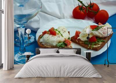 Poached eggs on sandwich of rye bread with sauce, tomatoes and arugula in white plate on blue background. Wall mural