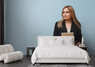 Blonde young business woman wearing black suit over blue plain background, carrying laptop Wall mural