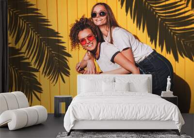 Candid shot of two lovely hipster women in glasses and denim clothes standing under the shadow of palm branches on a sunny summer day. Wall mural