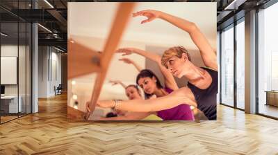 Beautiful adult women doing exercises on stretching ballet bar in Pilates class. Group of females doing yoga, pilates and fitness exercise indoors in studio. Wall mural