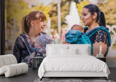 Two beautiful happy young mothers with baby carrier in park. Moms walking with infant in baby carrier. Happy mothers carrying her child by ergonomic baby carrier. Wall mural