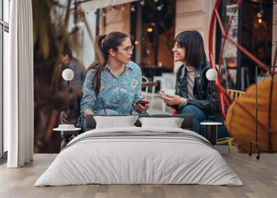 Two beautiful happywomen in cafe outdoors drinking coffee. Two female using mobile phone application and enjoying coffee outside in city Wall mural