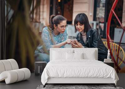 Two beautiful women in cafe outdoors looking at something on mobile phone. Two female using mobile phone application shopping online and enjoying coffee outside in city Wall mural