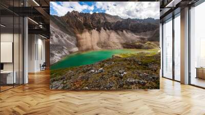 An elongated mountain lake with green water backlit by the sun through the clouds. White and brown mountain nearby. Rocky ground in the foreground. Stone talus. Horizontal. Wall mural