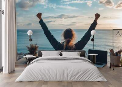 A woman sitting at her desk with arms raised in joy, celebrating success after embracing the corporate remote work lifestyle on an ocean-view balcony during sunrise. Wall mural