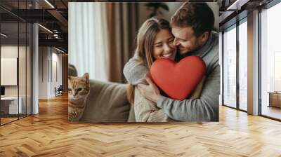 Happy young couple hugging and holding a red heart-shaped pillow at home on Valentine's Day, with a cat sitting nearby. Love concept. Wall mural