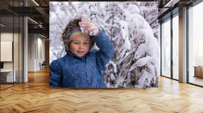 Little beautiful girl in winter clothes standing alone in the middle of a snowy forest. Girl in jacket with fur hood posing in winter forest. Shrubs and trees are covered with snow Wall mural