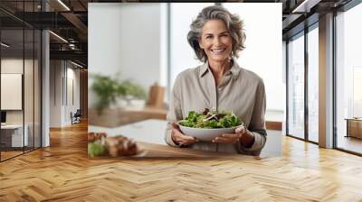 Adult Caucasian woman with short silver hair smiling while holding a bowl of fresh green salad in a bright modern kitchen. Wall mural