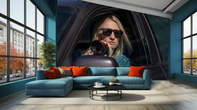 Young blonde woman in sunglasses takes pictures on a professional camera while sitting in a car. Concept of journalism, detective, papparation Wall mural