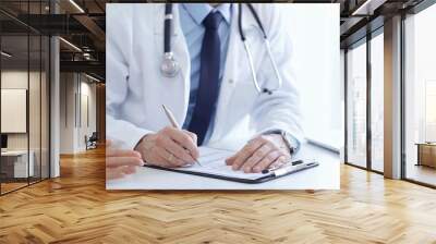 A doctor and a patient. The physician, wearing a white medical coat over a blue shirt and tie, is filling out a medical record form during a consultation in the clinic. Medical service Wall mural