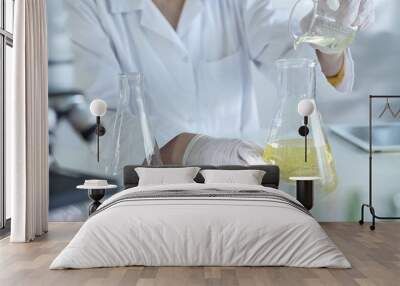 A scientist, wearing a lab coat and white protective gloves, is pouring a yellow oily liquid from one beaker to another near microscope in laboratory, close up. Medicine and science concept Wall mural