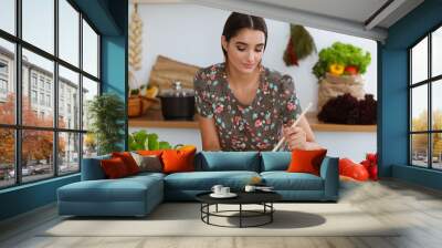 An attractive young dark-haired woman tastes a new recipe for a delicious salad mix while sitting and smiling in the kitchen. Cooking and householding concepts Wall mural