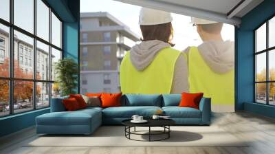 Man and woman architects or engineers wearing safety hard hats and vests holding blueprint and discussing something on construction site at sunrise, back view. Architecture and engineering Wall mural