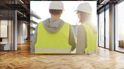 Two architects wearing safety hard hats and vests holding blueprint and discussing construction site project at sunrise, back view. Architecture, engineering and teamwork concept Wall mural