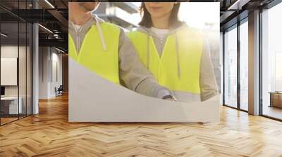 Two architects wearing safety hard hats and vests holding blueprint and discussing something on construction site at sunrise, front vertical view. Architecture engineering and teamwork concept Wall mural