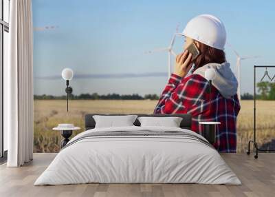 Woman engineer is talking on a smartphone in a field with wind turbines as the sun sets. Concept of clean energy and engineering audit Wall mural