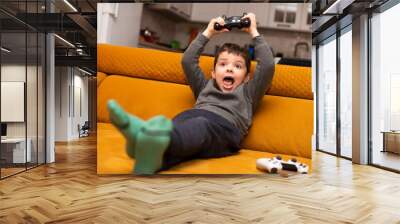 Cute 5 year old boy playing video game console while sitting on orange couch at home. Wall mural