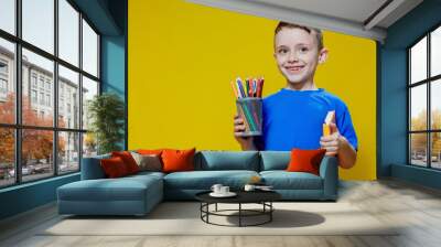 Smiling happy schooler in blue t-shirt holding multicolored pencils and book on yellow background Wall mural