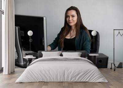 Brunette woman is working in front of a monitor in a office. Business woman working at computer at coworking space. Portrait of happy lady typing at workplace. Female professional working with pc. Wall mural