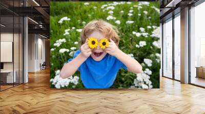 Funny Boy Kid and Daisies. Happy little blond hair child with flowers eyes laying on the grass with daisies flowers. Child with daisy eyes, on green grass in a summer park. Wall mural