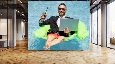 Funny business man in a business suit floating in the water in the pool. Remote work. Crazy freelancer. Business and summer. Business man drink summer cocktail and using laptop in pool. Wall mural