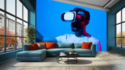 Handsome Bearded man in white t-shirt and VR headset enjoying experience playing a game and gesturing while standing against wall in neon light. Wall mural