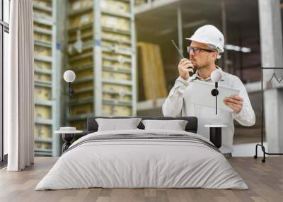 Male head engineer wearing white safety hardhat with walkie talkie and tablet inspecting construction site. Wall mural