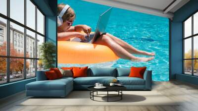 Little business man relaxing in the pool with laptop. Kid online working on laptop, swimming in a sunny turquoise water pool. Wall mural