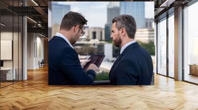 Offering deal, discussing new project. Photo of business partners talk conversation outdoor. Two handsome businessmen in suits discuss a working project against the background of urban city. Wall mural