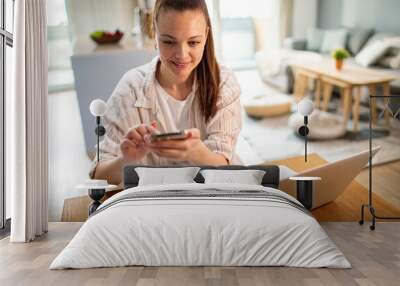 Young female freelancer working on laptop using smartphone at home Wall mural