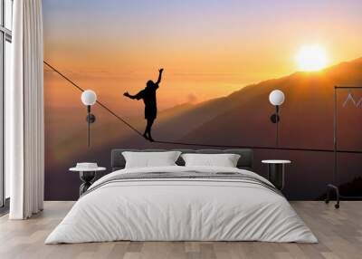 Silhouette of young man balancing on slackline high above clouds and mountains. Slackliner balancing on tightrope during sunset, highline silhouette. Wall mural