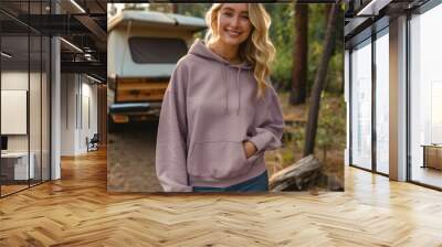 A smiling young woman in a hoodie stands outdoors in front of a camper van surrounded by a forest setting Wall mural