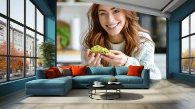 A young woman smiles holding a slice of avocado toast in a warmly lit home setting Wall mural