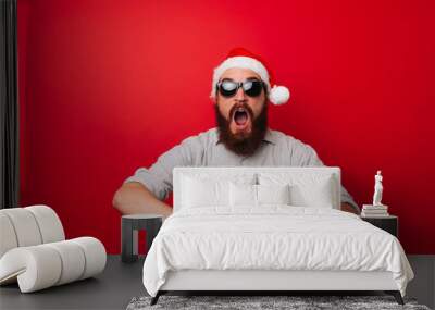 Amazed bearded man in santa claus hat pointing at smartwatch over red background Wall mural