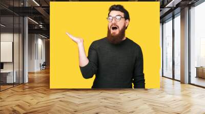 Amazed man with beard holding copyspace in palm standing over yellow background Wall mural