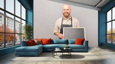 Bald smiling barista man is holding a blank clean black board for copy space. Studio shot over grey background. Wall mural