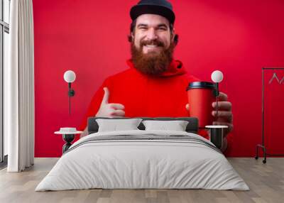 Bearded guy  in rd hoody with toothy smile holding a cup and showing thumb up over isolated red background Wall mural