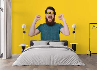 Bearded happy man with futuristic glasses making win gesture on yellow background. Wall mural